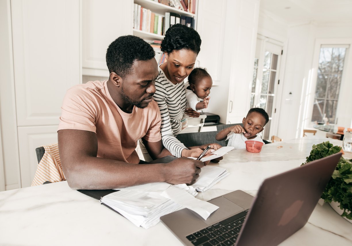 A man sits at a kitchen table with bills and a laptop, while a woman holding a baby stands beside him, and a young child eats at the table. The family appears to be reviewing finances together.