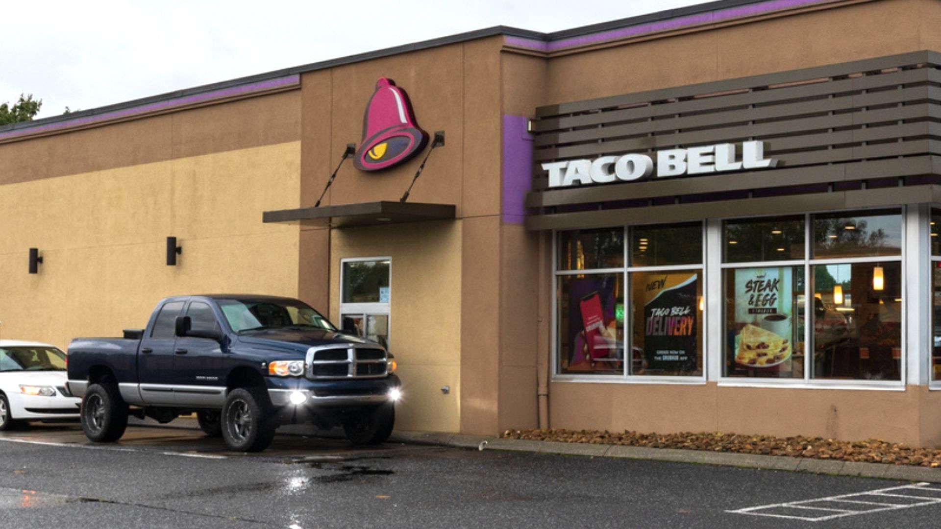 A dark blue pickup truck is parked in front of a Taco Bell restaurant on a rainy day. The restaurant has tan walls, large windows displaying menu items, and a prominent Taco Bell sign above the entrance.