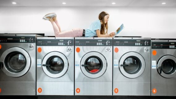 A young woman lies on top of a row of washing machines in a laundromat, reading a magazine and smiling. The machines are silver, numbered, and the setting is clean and modern.