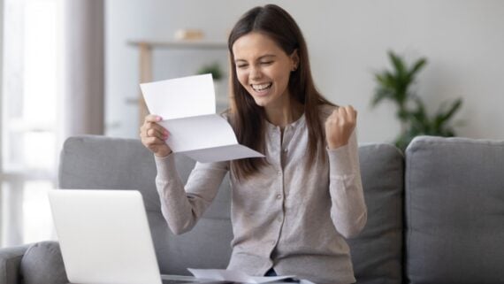A young woman sits on a couch in front of a laptop, smiling and raising her fist in excitement while holding and reading a letter, suggesting she has received good news.
