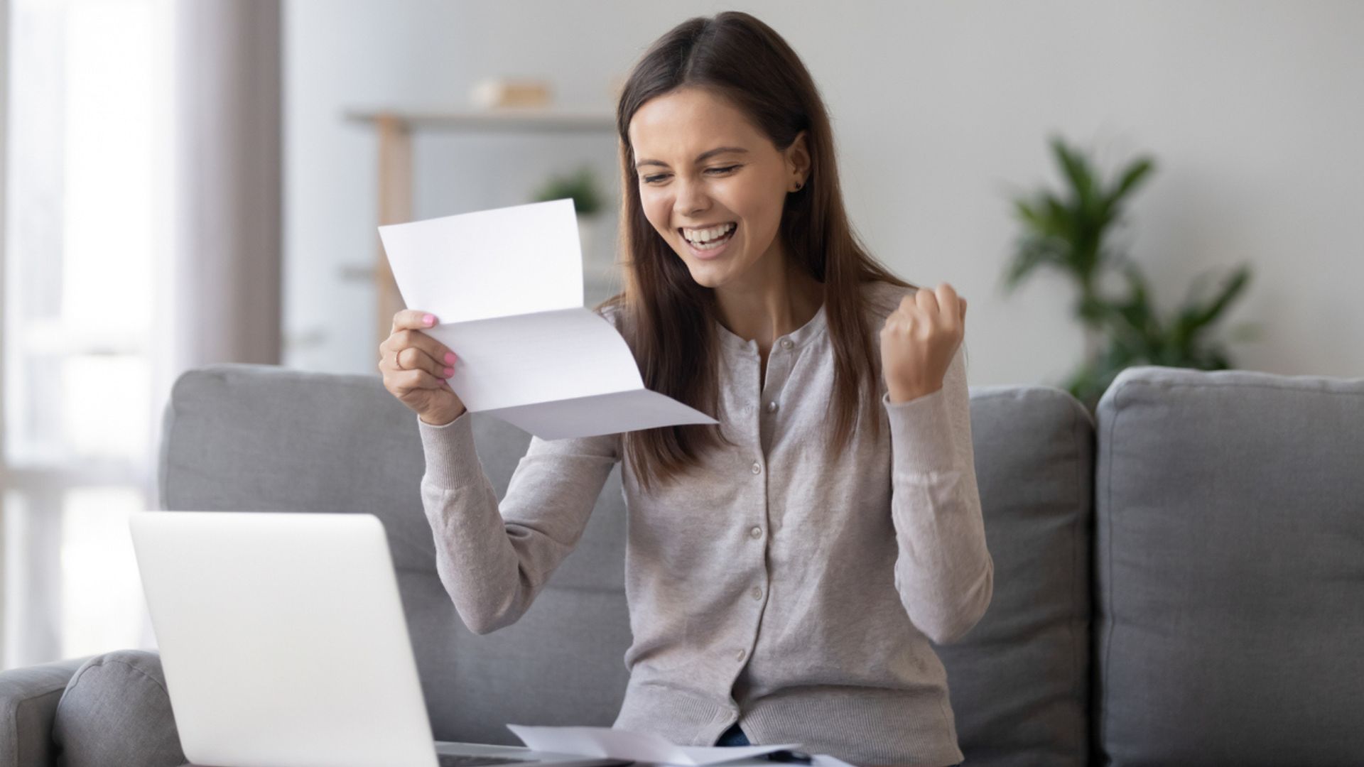 A young woman sits on a couch in front of a laptop, smiling and raising her fist in excitement while holding and reading a letter, suggesting she has received good news.
