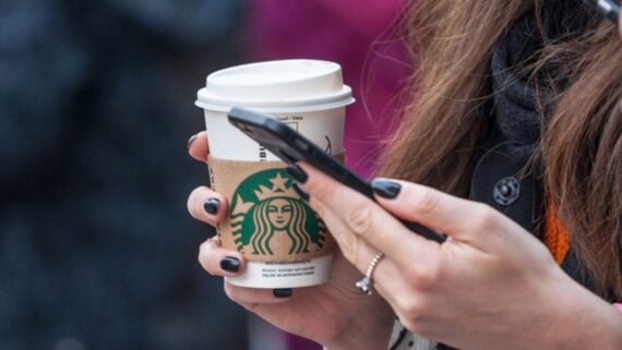 A person with black nail polish holds a Starbucks coffee cup in one hand and a smartphone in the other, with only their hands visible and a blurred background.