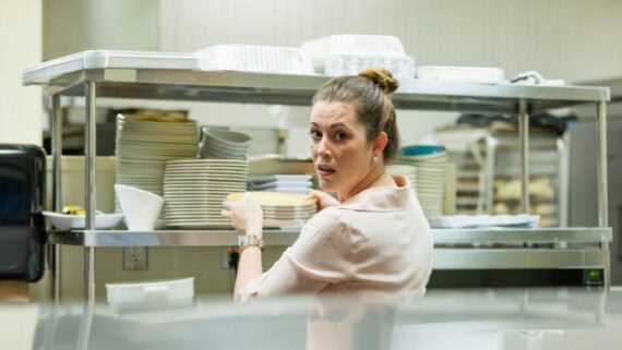 A woman in a light pink blouse stands in a commercial kitchen, holding a stack of plates and looking over her shoulder toward the camera. Shelves filled with more dishes and trays are visible behind her.