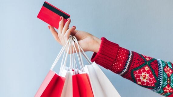 A person wearing a red holiday sweater holds several red and white shopping bags in one hand and a red credit card in the other, against a light gray background.