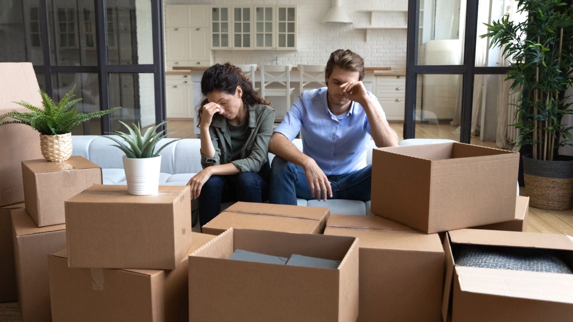 A stressed couple sits on a couch surrounded by unpacked moving boxes in a modern living room, both with heads in their hands. Houseplants and kitchen cabinets are visible in the background.