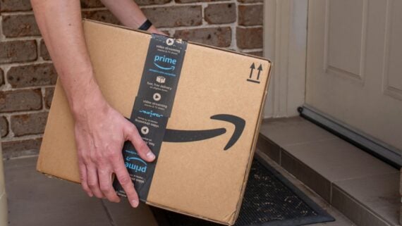 A person places a large Amazon Prime package on a doormat outside a front door, with a brick wall in the background.