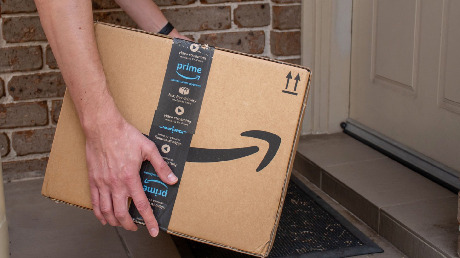 A person places a large Amazon Prime package on a doormat outside a front door, with a brick wall in the background.