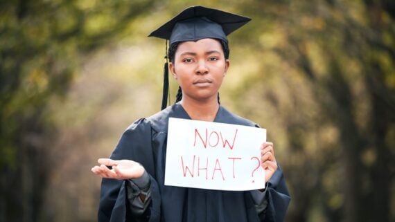 A graduate in a cap and gown stands outdoors, holding a sign that reads "NOW WHAT?" and gesturing with one hand, looking uncertain. The background is blurred with green foliage.