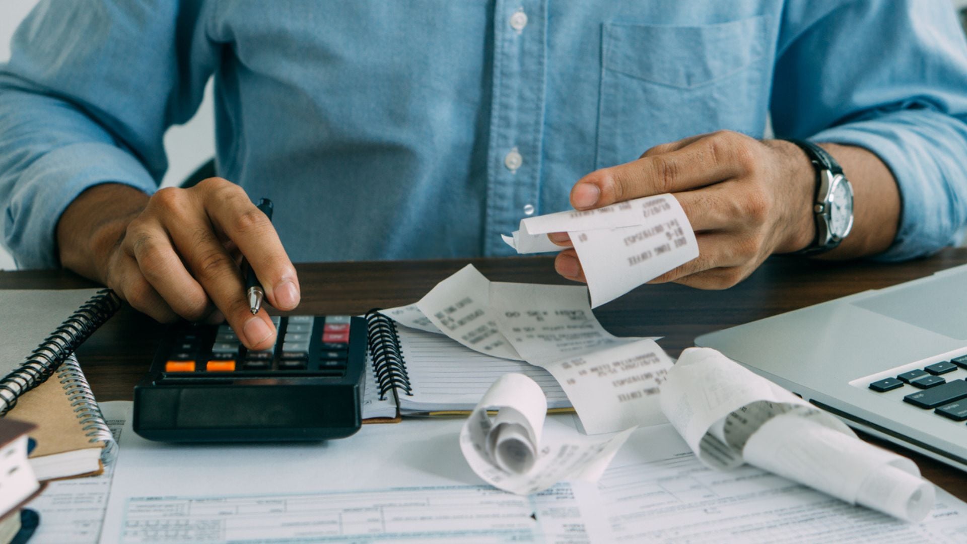 A person in a blue shirt uses a calculator and holds several receipts, surrounded by paperwork, notebooks, and a laptop on a desk, suggesting budgeting or financial calculations.