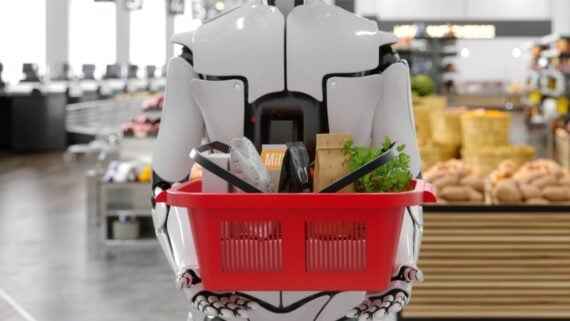 A humanoid robot holding a red shopping basket filled with groceries in a supermarket, with shelves of bread and produce in the background.