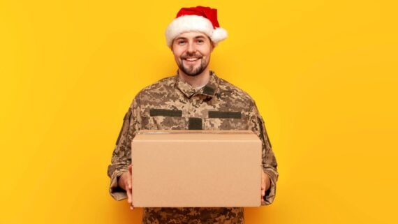 A smiling person in military uniform and a Santa hat holds a cardboard box against a bright yellow background.