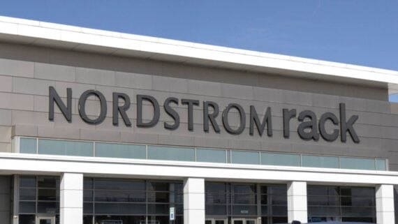 The exterior of a Nordstrom Rack store, featuring large black letters spelling “NORDSTROM rack” on a light gray building facade under a clear blue sky.