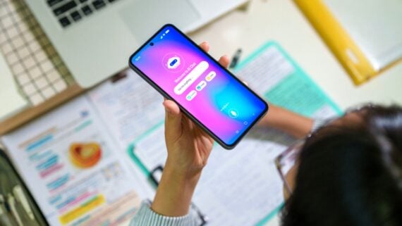 A person holds a smartphone displaying a language learning app; notebooks, papers with handwritten notes, and part of a laptop are visible on a desk beneath them.