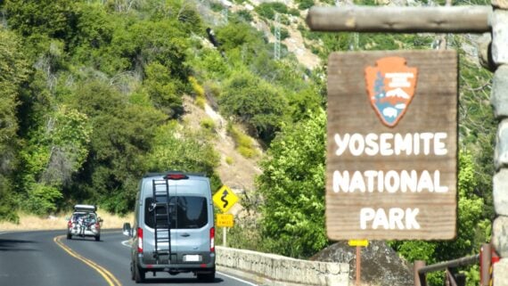 A van and a car tow a trailer on a winding road through a forested area; a wooden sign in the foreground reads "Yosemite National Park" beside the road.