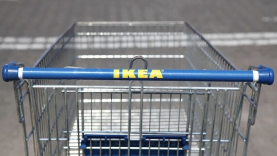 A close-up view of an empty metal shopping cart with a blue handle that has the yellow IKEA logo, photographed outdoors on a paved surface.