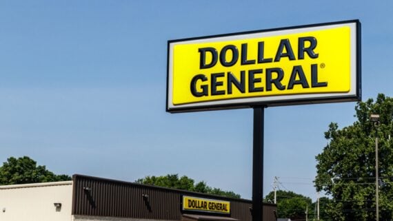 A large yellow Dollar General sign on a tall pole stands in front of a Dollar General store, with trees and a clear blue sky in the background.