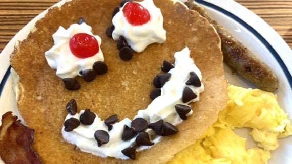 A pancake decorated with whipped cream, cherries, and chocolate chips to form a smiling face, served with scrambled eggs, a sausage link, and a strip of bacon on a white plate.