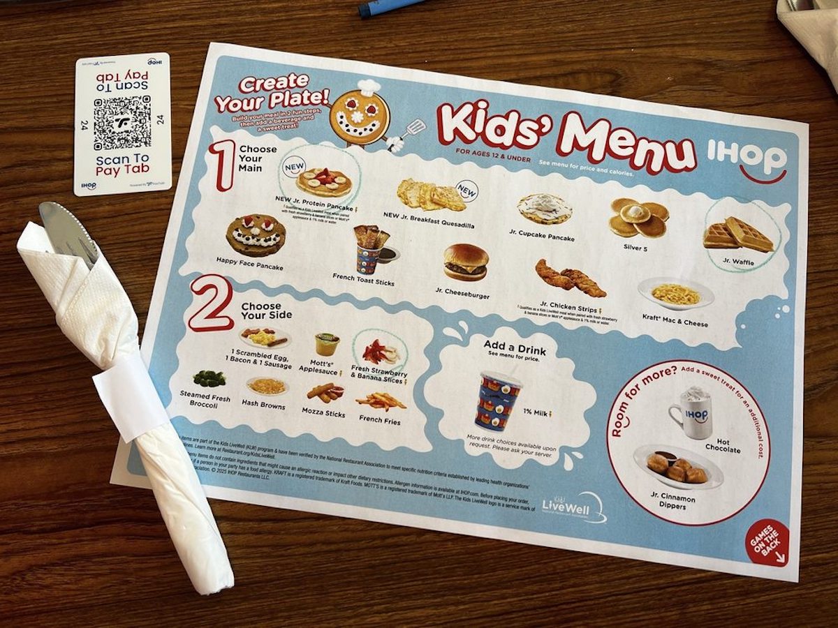 A colorful IHOP kids’ menu on a wooden table shows meal options with images, including pancakes, eggs, cheeseburgers, and sides. A wrapped napkin with utensils and a QR code to pay the tab are also visible.