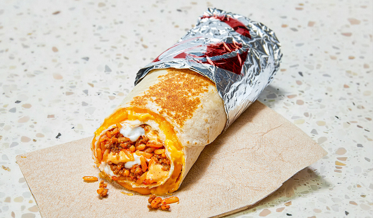 A Taco Bell Quesarito burrito wrapped in foil and partially unwrapped sits on a napkin. The filling includes rice, ground meat, cheese, and sauce, all wrapped in a toasted tortilla. The background is a speckled, light-colored surface.