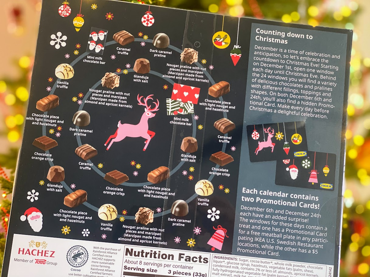 A festive advent calendar featuring chocolates arranged in a circle around a pink reindeer, with Christmas-themed icons and instructions for counting down to Christmas. A blurred decorated Christmas tree is in the background.
