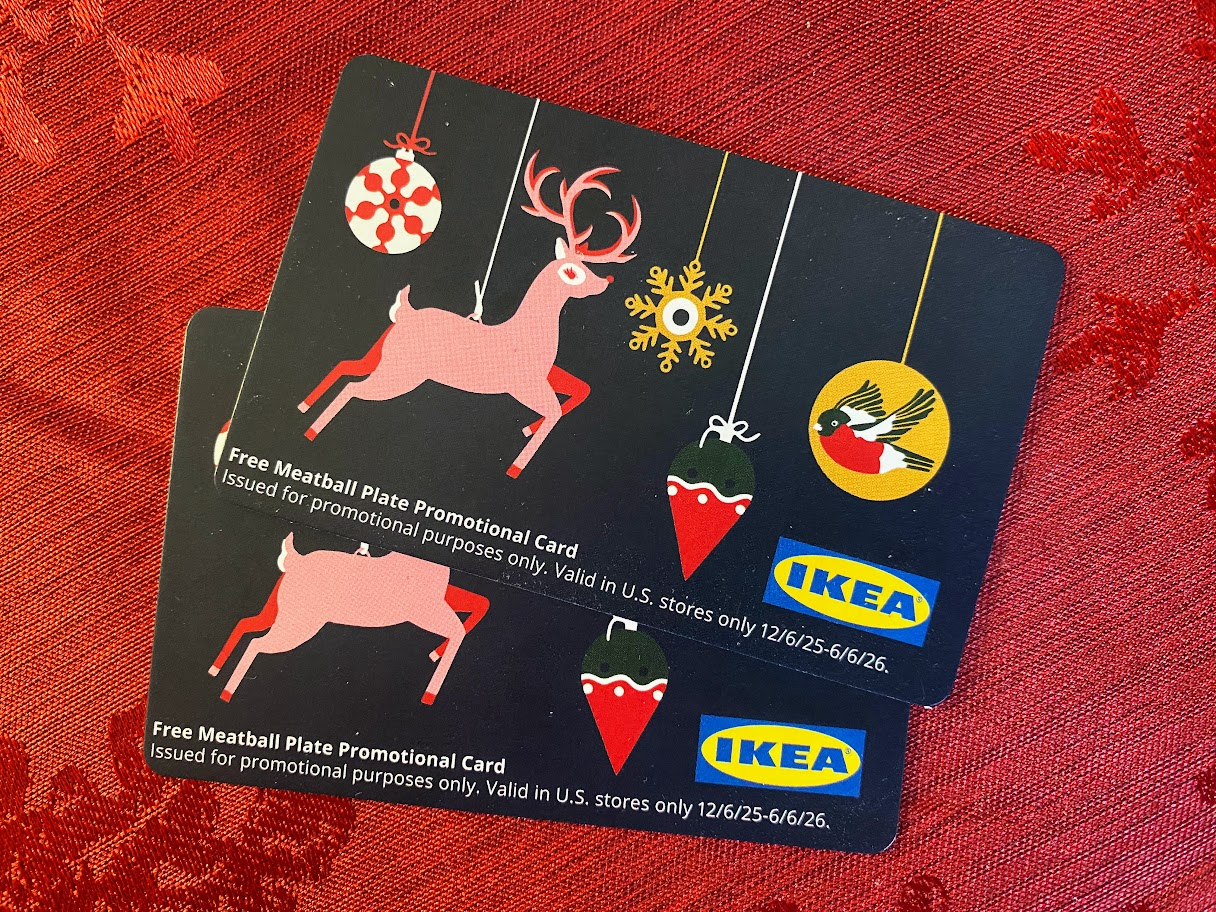 Two IKEA promotional cards featuring a festive design with a reindeer, ornaments, bird, and hearts on a black background. The cards offer a free meatball plate and are set on a red textured surface.