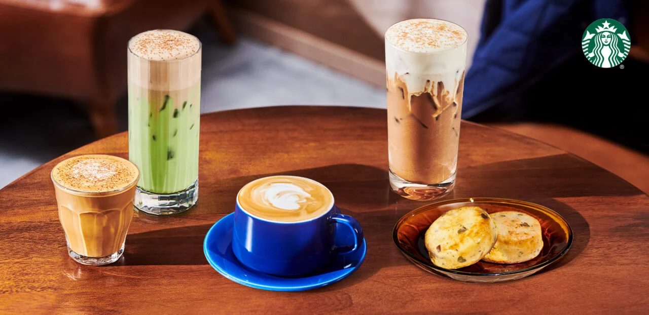 Four drinks and a plate of two snacks are displayed on a wooden table, featuring lattes and iced beverages in glasses and a blue cup. The Starbucks logo hints at their new fast-food menu in the top right corner.