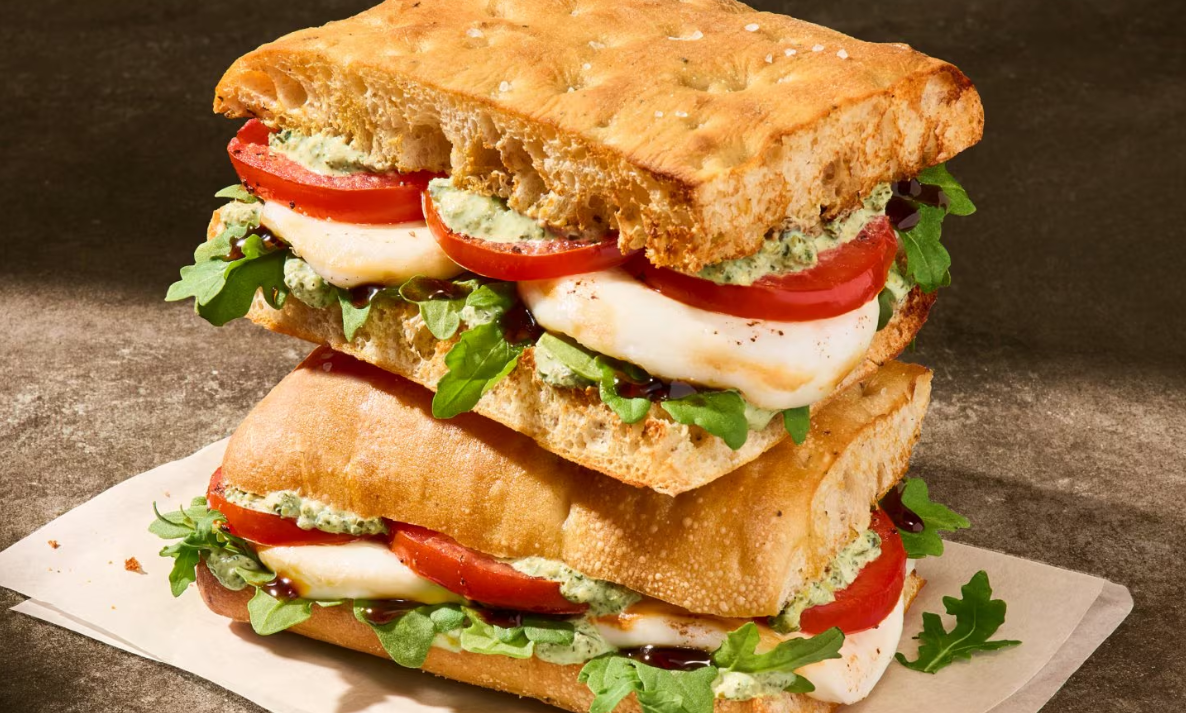 Two stacked ciabatta sandwiches from our new fast-food menu, filled with fresh mozzarella, tomato slices, arugula, pesto spread, and a drizzle of balsamic glaze, placed on parchment paper against a brown background. Toasted Caprese focaccia Panera