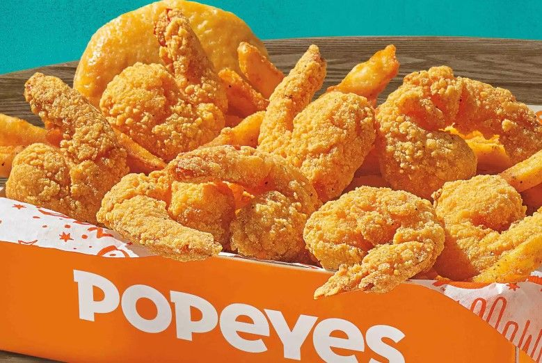 A box of crispy fried shrimp and a biscuit from Popeyes, part of their new fast-food menu, served in branded orange packaging on a wooden surface with a blue background.