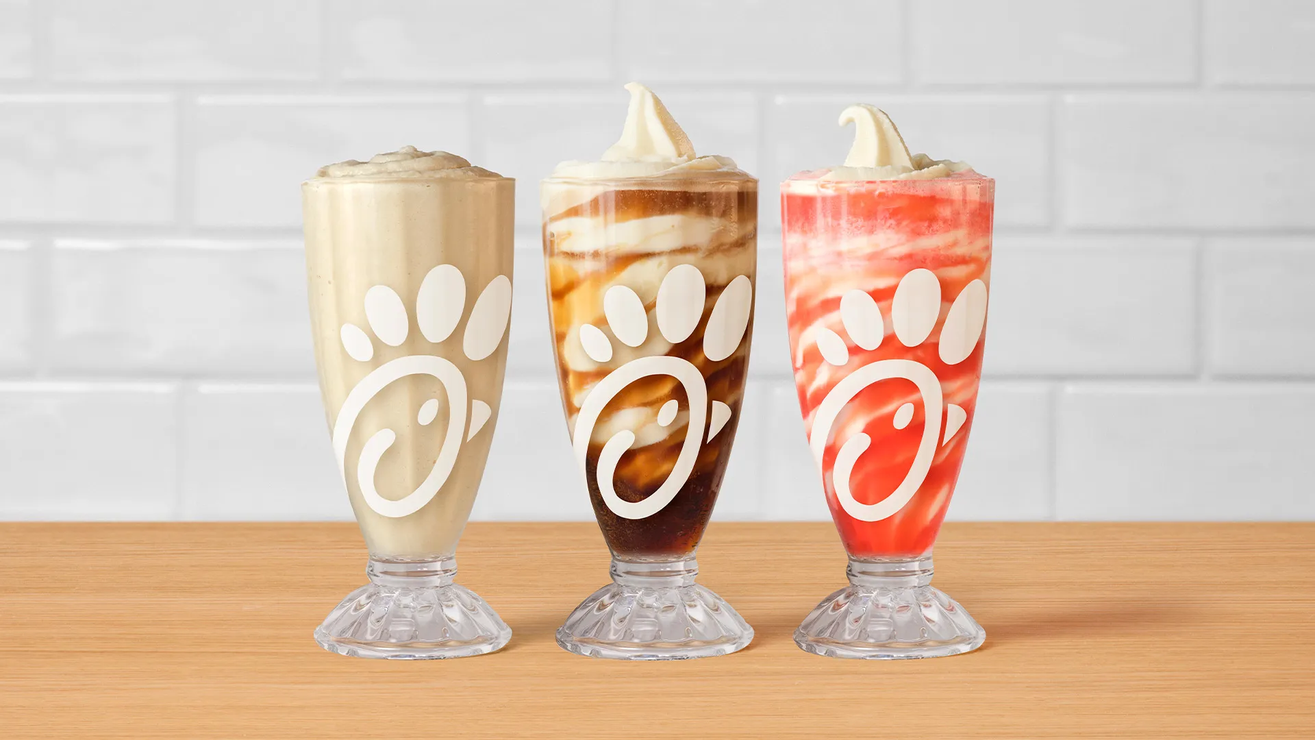 Three tall glasses of milkshake from the new fast-food menu, topped with soft serve ice cream and featuring caramel, chocolate, and strawberry swirls, are arranged on a wooden surface against a white tile background.