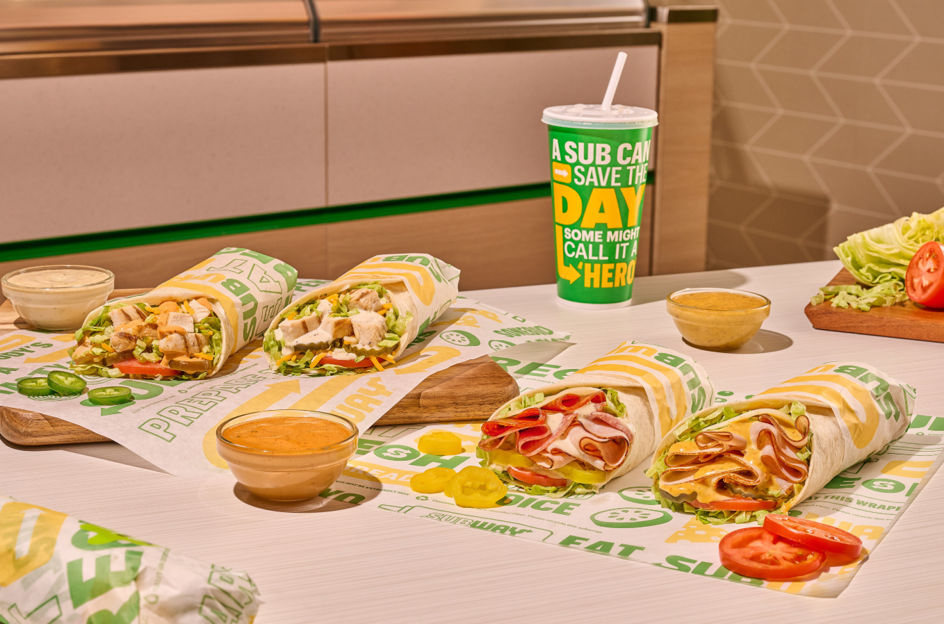 Three sub sandwiches from the new fast-food menu are cut in half and filled with meats and vegetables, displayed on wrappers with sliced tomatoes, jalapeños, lettuce, yellow peppers, sauces, and a paper cup with a straw on a white table.