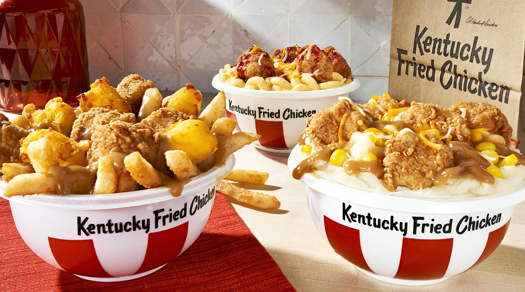 Three KFC bowls from the new fast-food menu are filled with fried chicken, mashed potatoes, corn, cheese, fries, and gravy. A brown Kentucky Fried Chicken bag and a red container sit in the background.