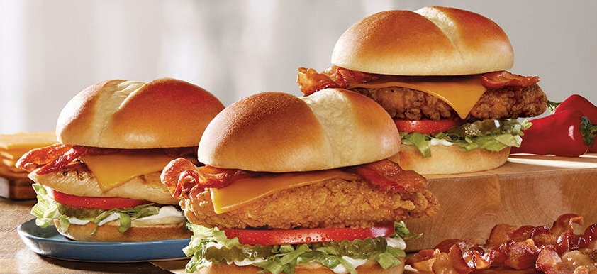 Three crispy chicken sandwiches from the new fast-food menu are topped with bacon, cheddar cheese, lettuce, tomato, and mayonnaise on fresh buns. Extra bacon strips sit in the corner, all set on a rustic wooden surface.