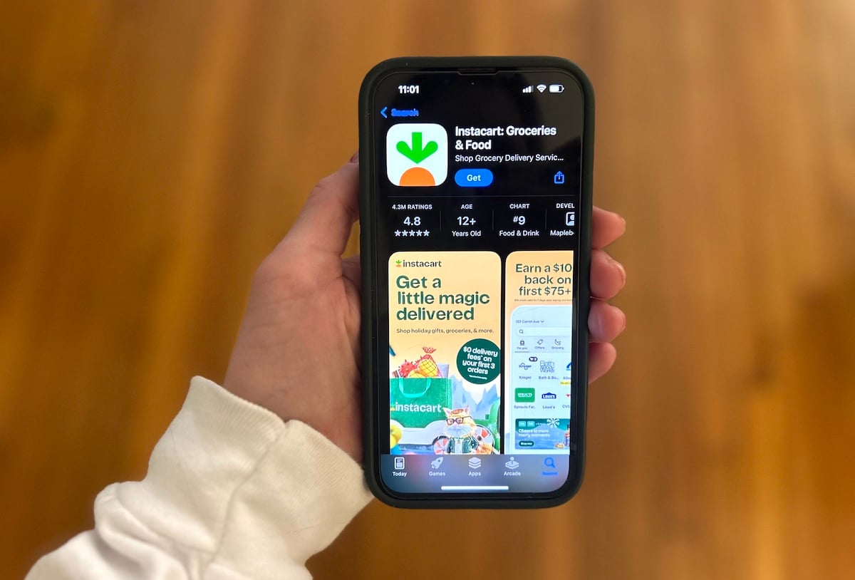 A person holds a smartphone displaying the Instacart app page in the App Store, showing the app's logo, rating, and promotional banner. The background is a blurred wooden floor.
