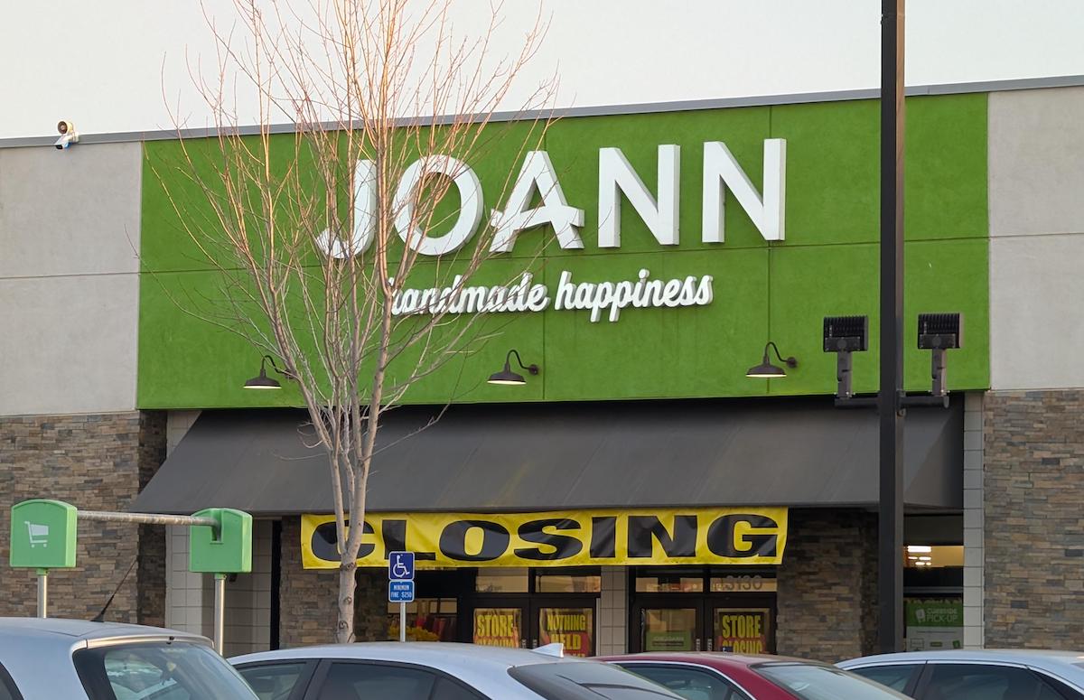 The front of a JOANN fabric and craft store with a large green sign, and yellow “CLOSING” banners hanging in the windows. Several parked cars are visible in front of the building.
