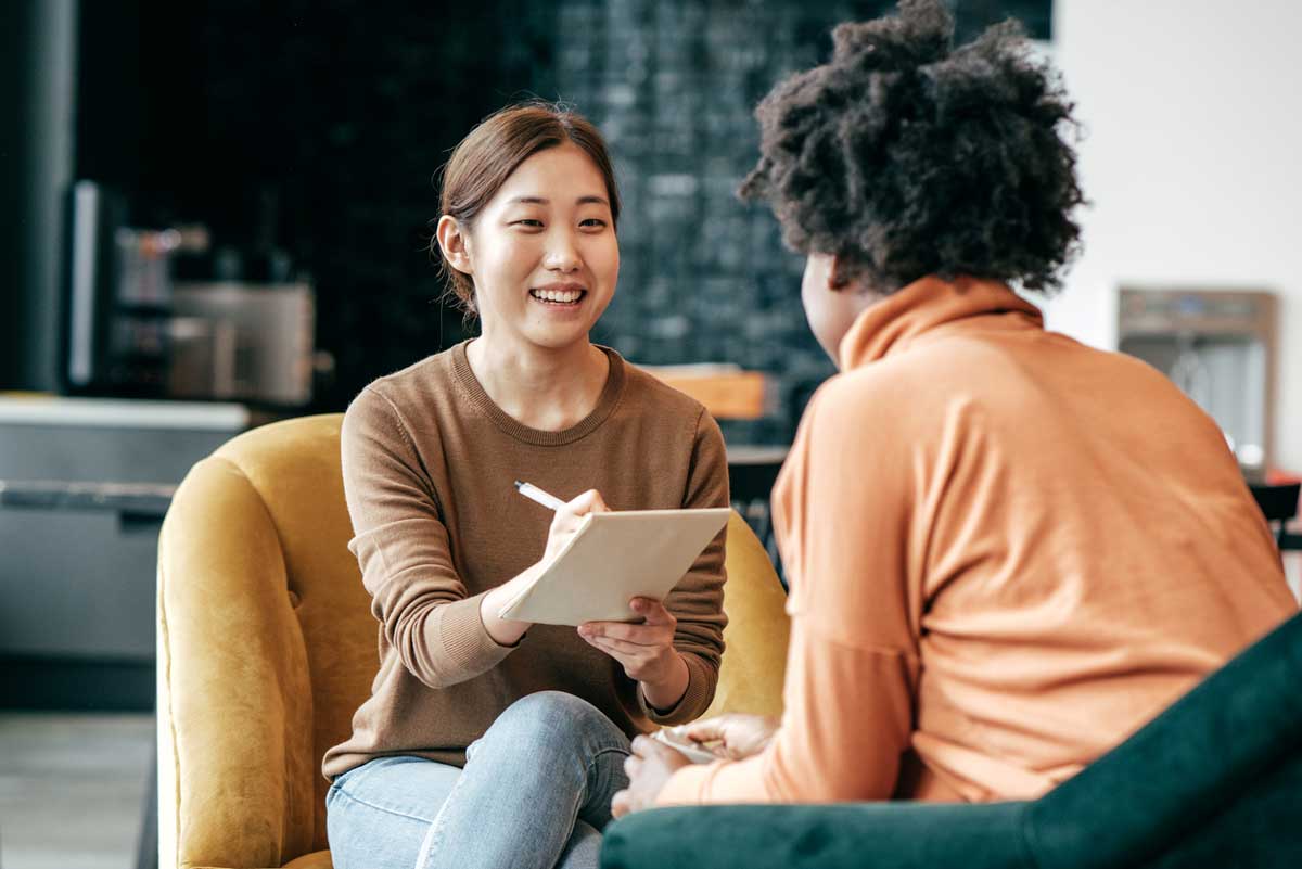 Two women sit facing each other in armchairs, smiling and talking. One holds a clipboard, appearing to take notes or ask questions about how to renegotiate bills in January, in a bright, modern indoor space.