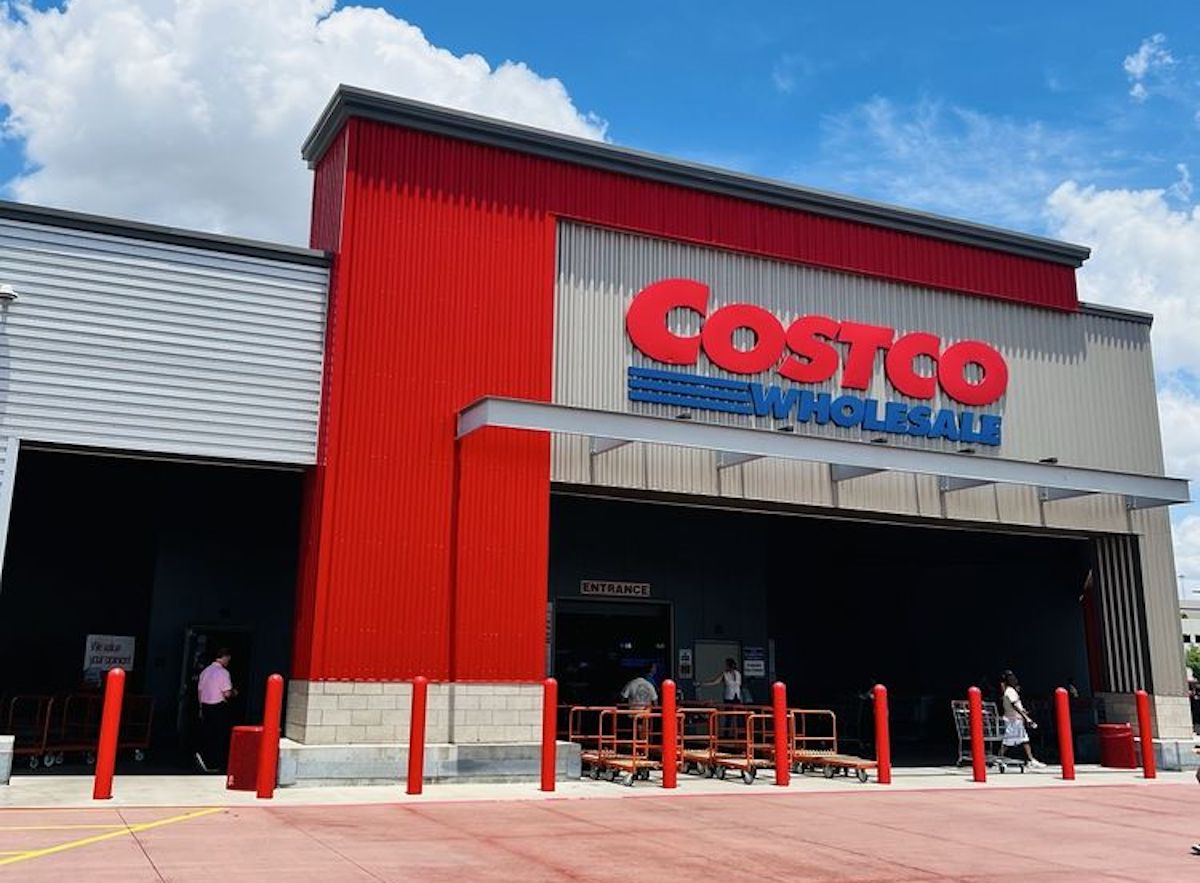 The image shows the exterior of a Costco Wholesale store with a red and gray facade, large red and blue signage, and people entering or exiting near shopping carts under a partly cloudy sky.