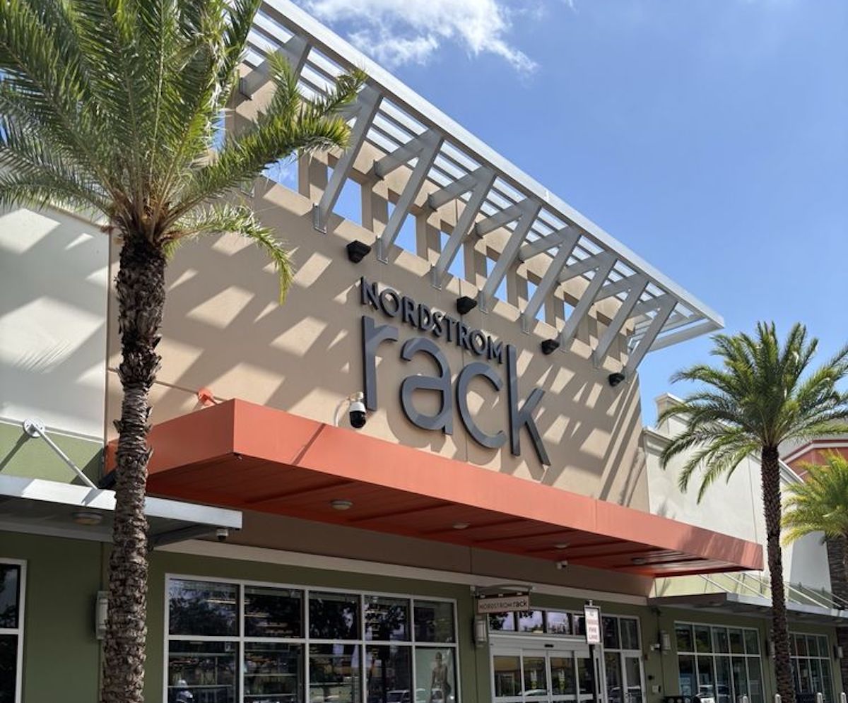 The exterior of a Nordstrom Rack store with large signage, palm trees, and a partly cloudy blue sky overhead. The building features modern architectural elements and a red awning above the entrance.