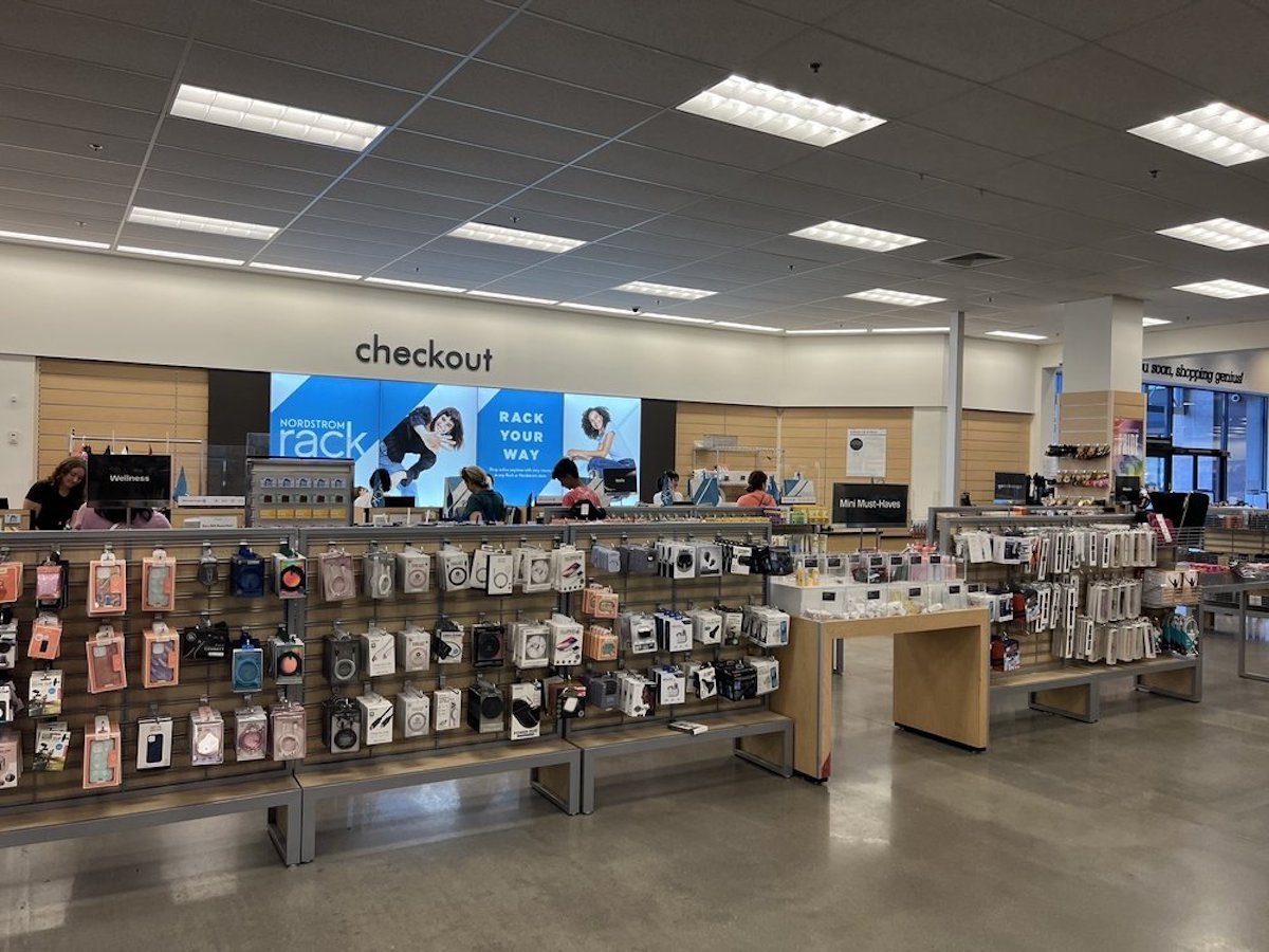 A retail store interior shows a checkout counter with staff and customers. In the foreground, shelves display electronics and accessories. A large sign above the counter reads "checkout" and advertising panels are visible behind.