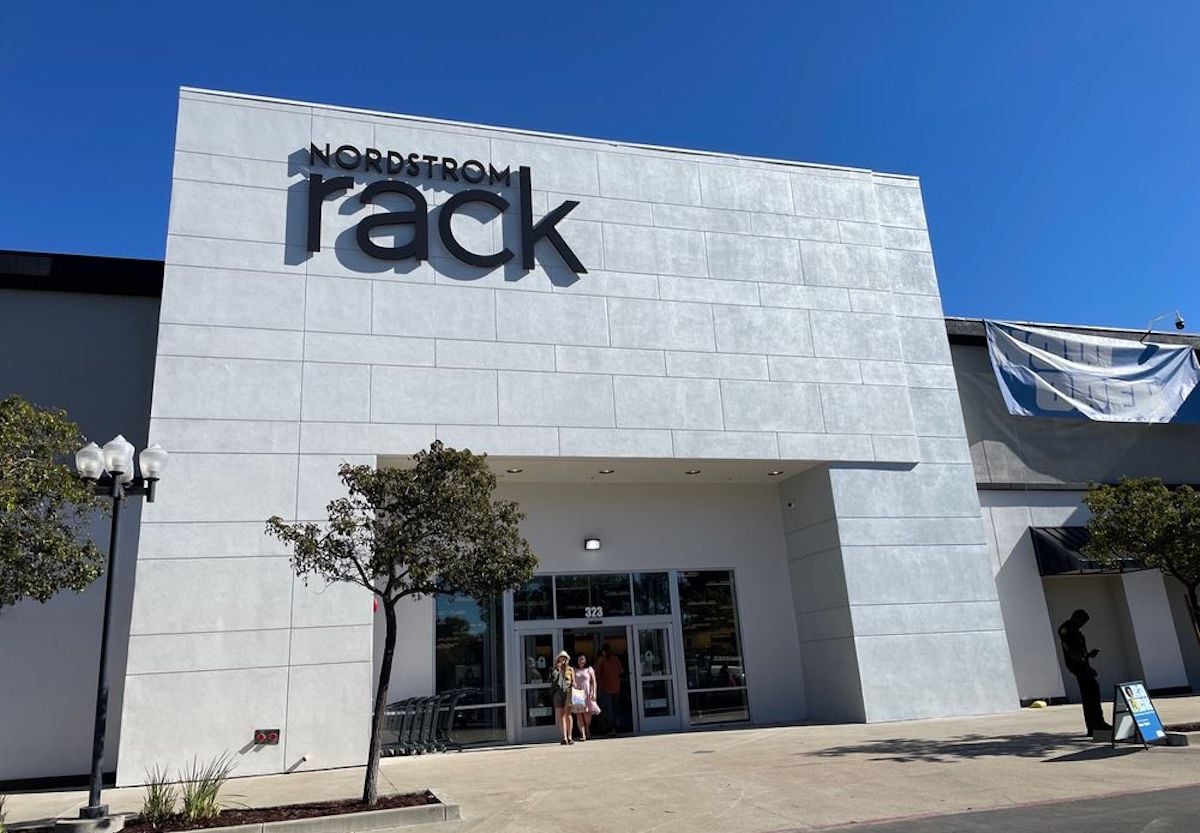 The exterior of a Nordstrom Rack store, with large signage on a light gray building under a clear blue sky; two people stand near the entrance, and trees are visible in front of the store.