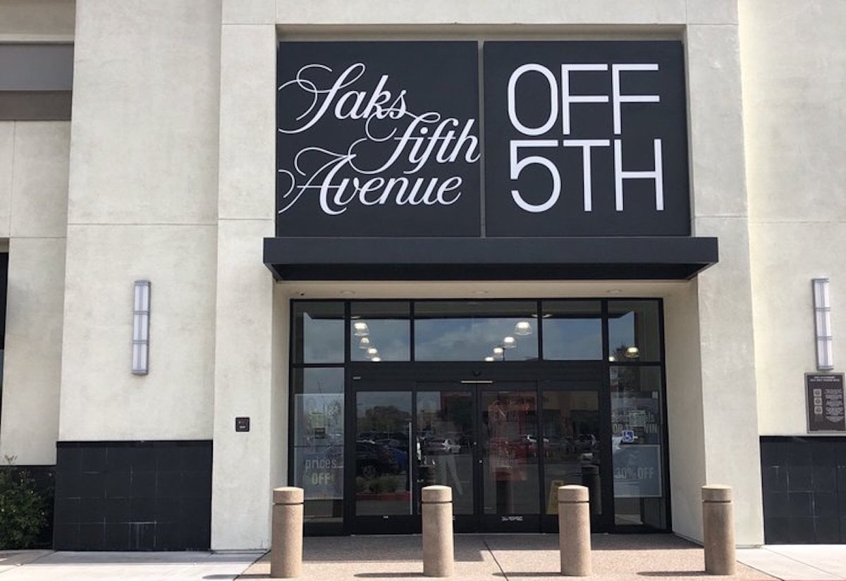 The entrance of a Saks Fifth Avenue OFF 5TH store, featuring large black and white signage above glass doors, with bollards in front and reflections visible in the windows.