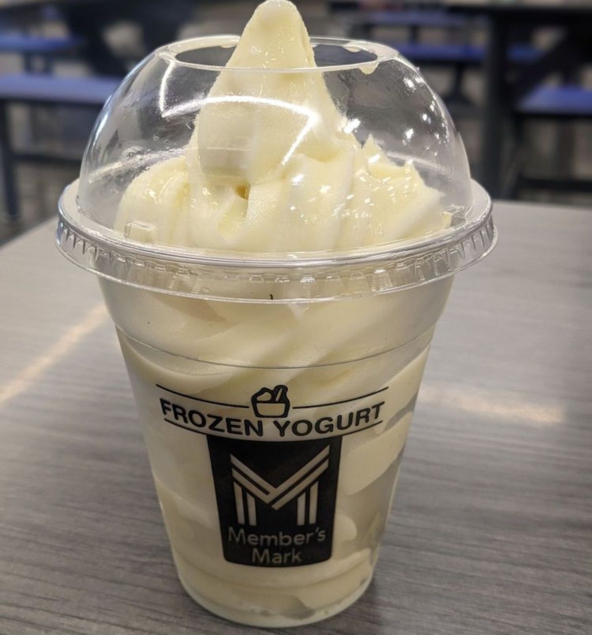A clear plastic cup of vanilla frozen yogurt with a dome lid sits on a gray tabletop. The cup is labeled "FROZEN YOGURT Member's Mark." Blue chairs and tables are visible in the blurred background.