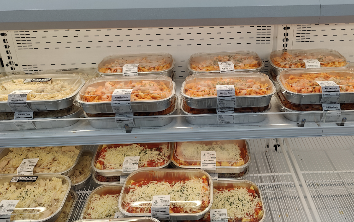 Several shelves in a grocery store display trays of prepared meals, including pasta and lasagna, covered with clear plastic lids and labeled with ingredient and pricing information.