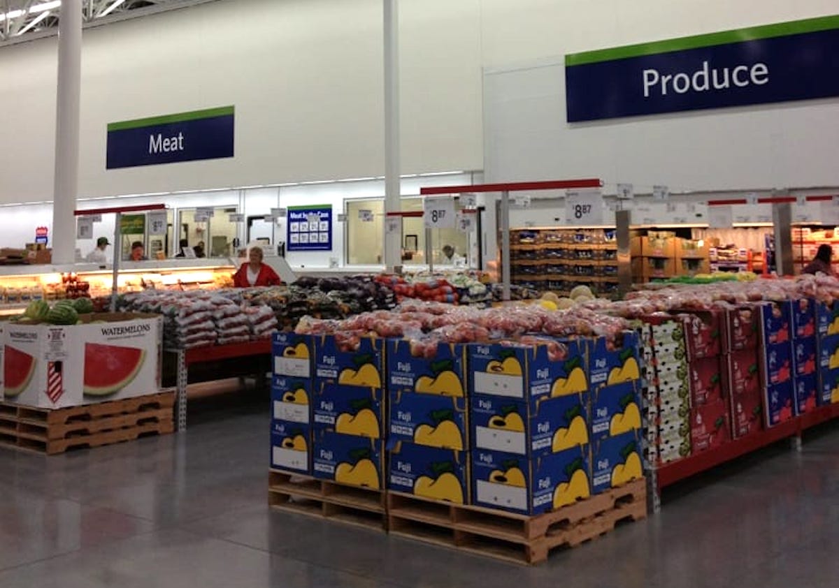 A grocery store interior displays fresh produce like apples and watermelons stacked on pallets under a “Produce” sign, with the “Meat” section in the background. Shoppers and workers are visible.