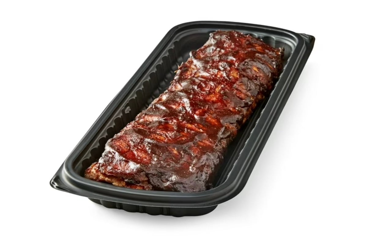 A rack of glazed, cooked ribs sits in a black plastic tray against a white background. The ribs have a shiny, caramelized surface, indicating they are covered in barbecue sauce.