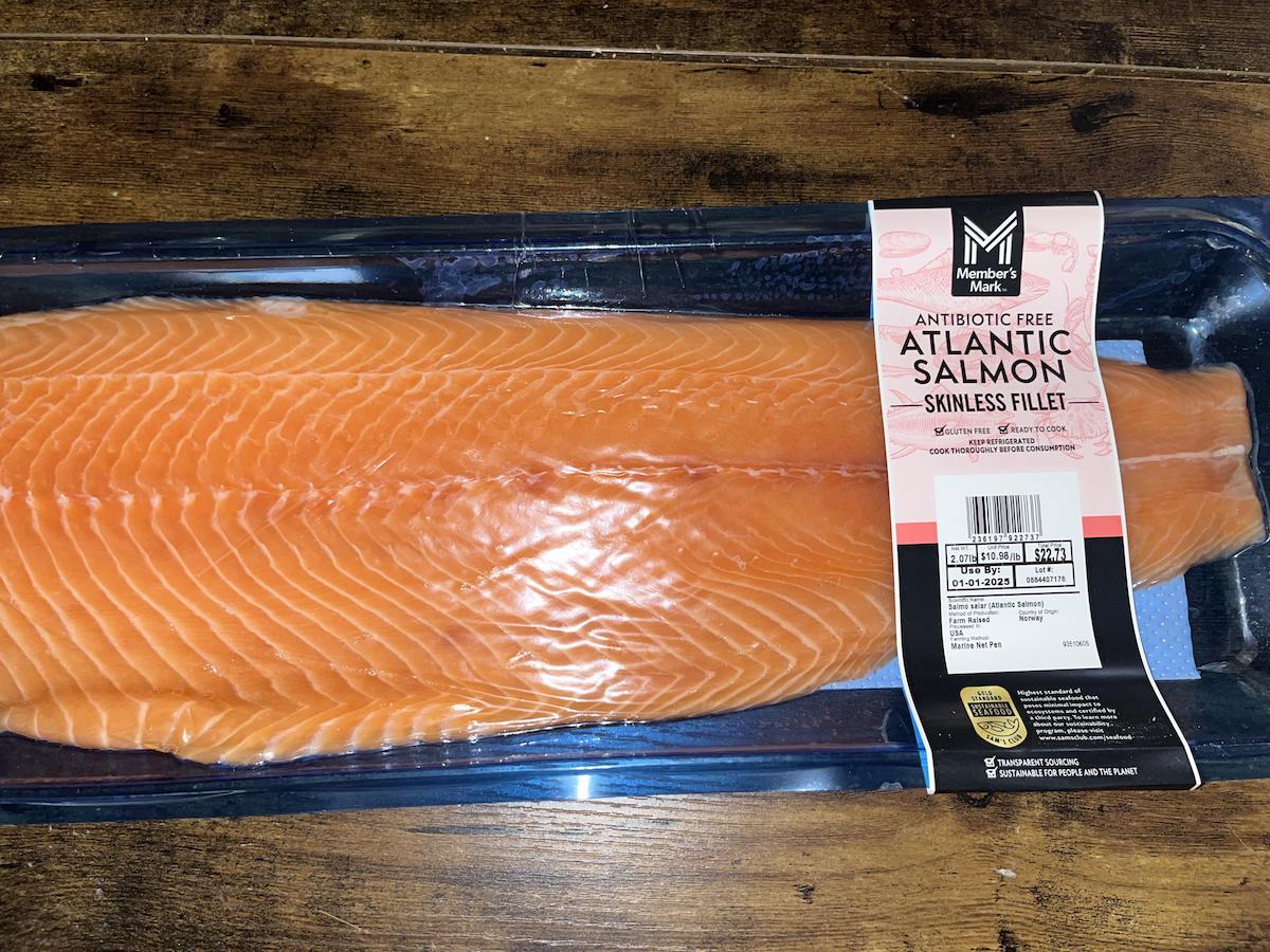 A package of Member's Mark antibiotic-free Atlantic salmon skinless fillet sits on a wooden surface. The label shows nutritional information and expiration date. The salmon fillet is orange-pink and placed in a clear plastic tray.