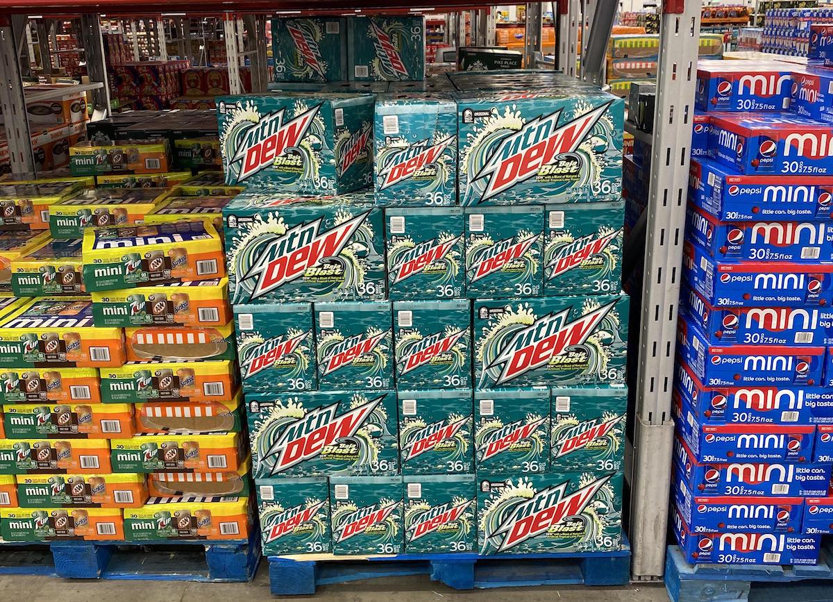 Stacks of Mountain Dew 36-pack boxes are displayed on a blue pallet in a store, surrounded by boxes of various mini snack packs and beverages on both sides.
