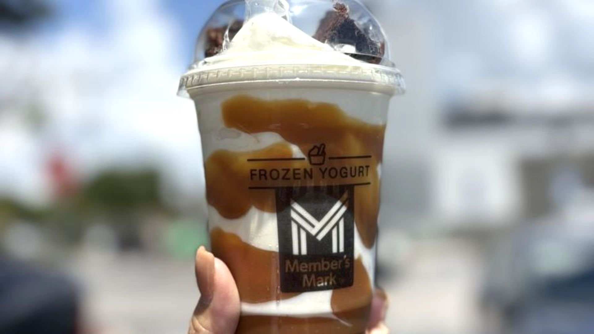 A hand holds a clear cup of Member’s Mark frozen yogurt topped with caramel sauce, brownie pieces, and a dome lid. The background is blurred with hints of blue sky and buildings.