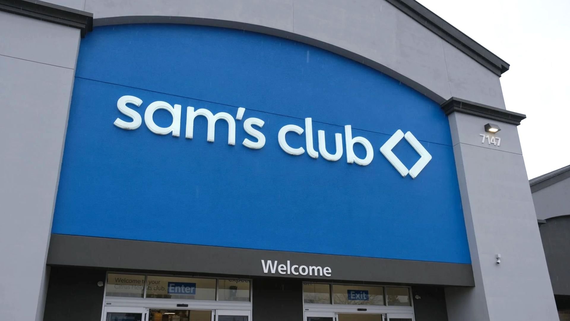 The front entrance of a Sam's Club store, featuring a large blue sign with the Sam's Club logo above the doors and the word "Welcome" visible below.