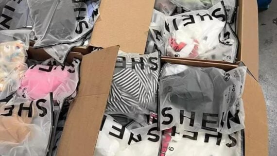 Open cardboard boxes filled with clear plastic bags of folded clothing from SHEIN, displaying various colors and patterns like stripes, pink, and white.
