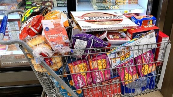 A grocery cart filled with frozen pizzas, breakfast pastries, bread, pepperoni, chips, and other packaged foods, in front of a freezer section at a grocery store.